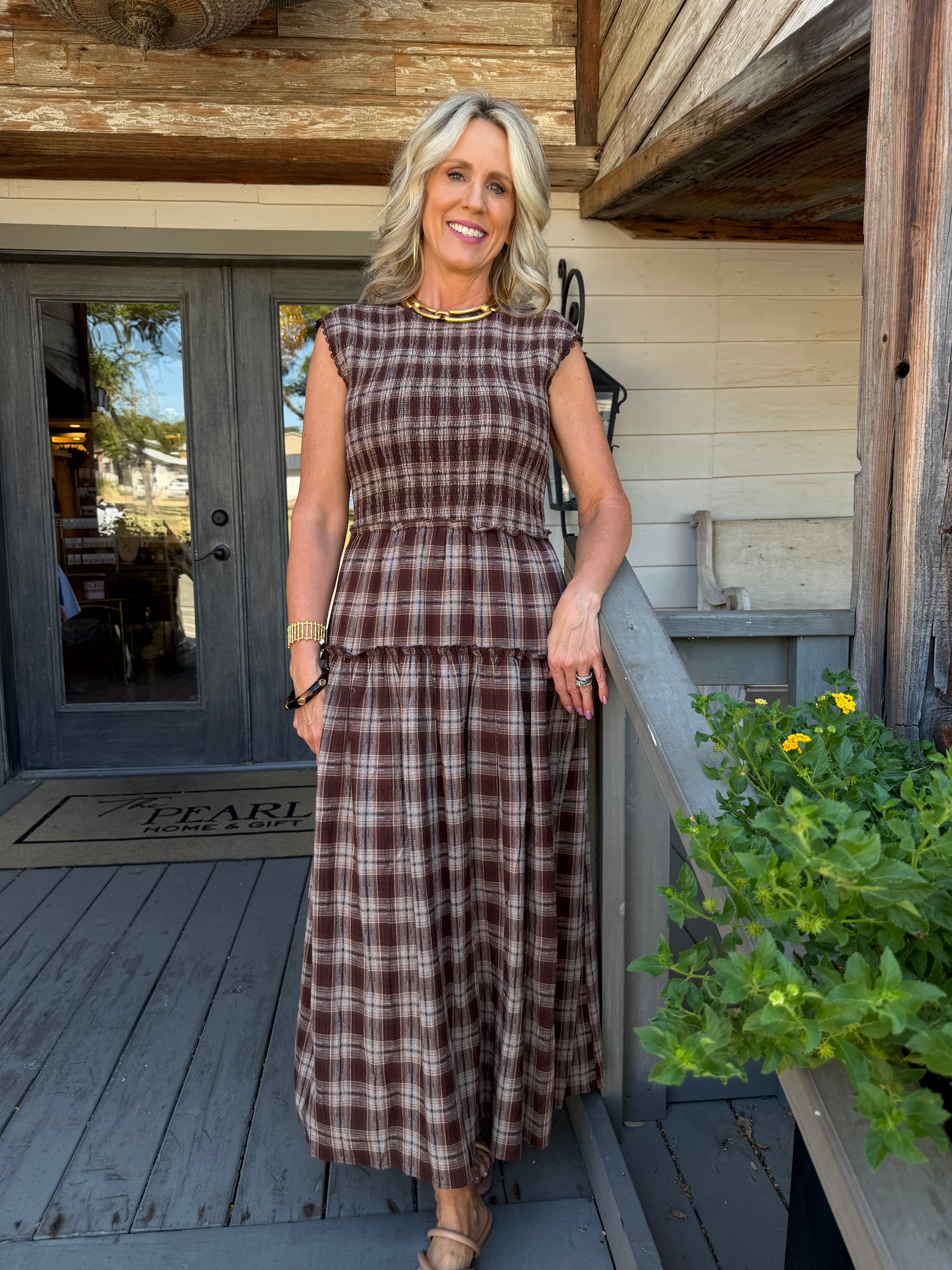 A woman stands on a rustic wooden porch wearing a brown and beige plaid sleeveless maxi dress with a tiered waist. The background features weathered wood siding, a gray door, and a potted green plant with yellow flowers.