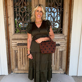 A woman stands in front of a rustic wooden door with decorative metal grates, wearing an olive green tiered midi dress with ruffled short sleeves and holding a brown polka-dot handbag.