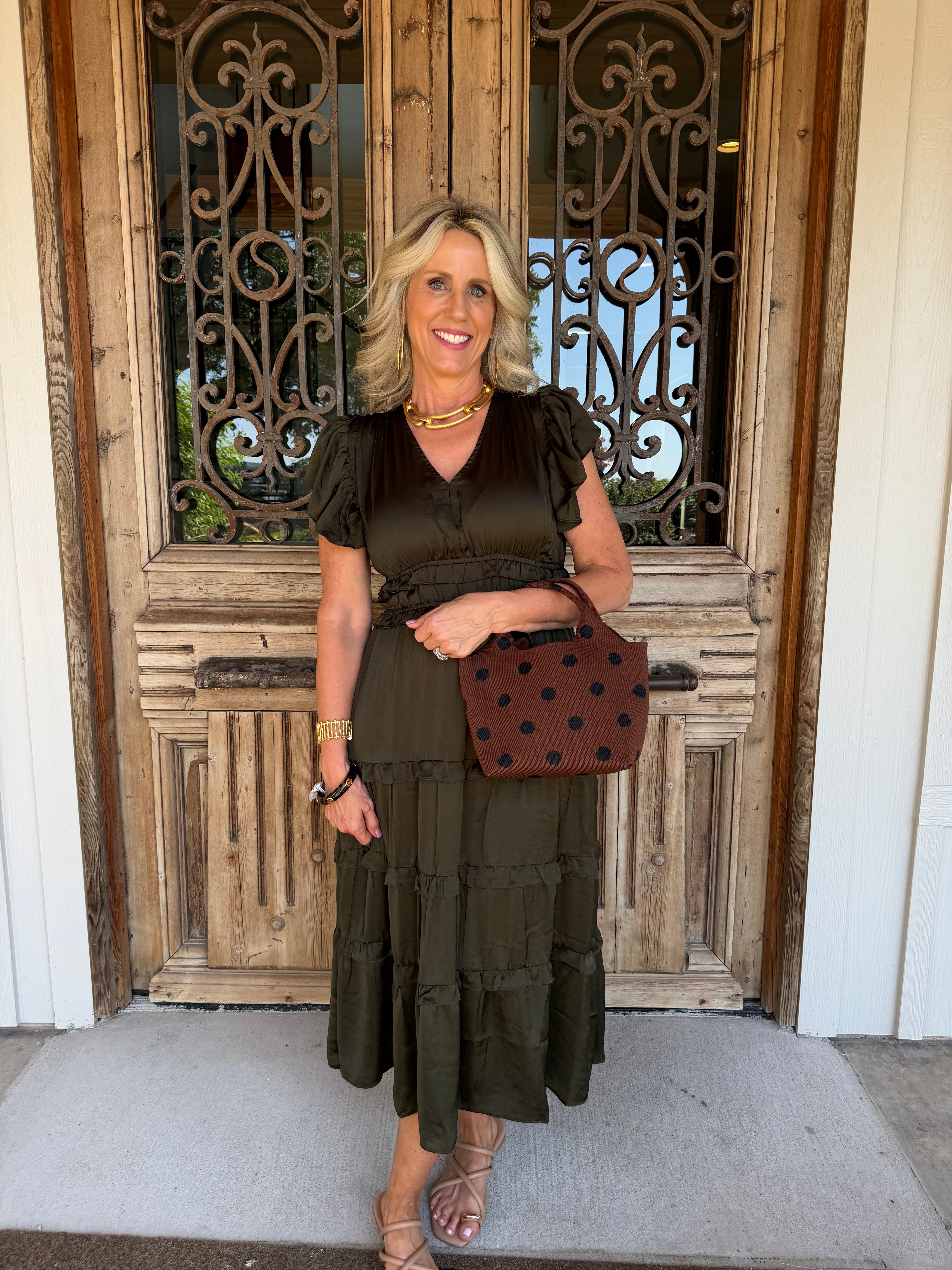 A woman stands in front of a rustic wooden door with decorative metal grates, wearing an olive green tiered midi dress with ruffled short sleeves and holding a brown polka-dot handbag.