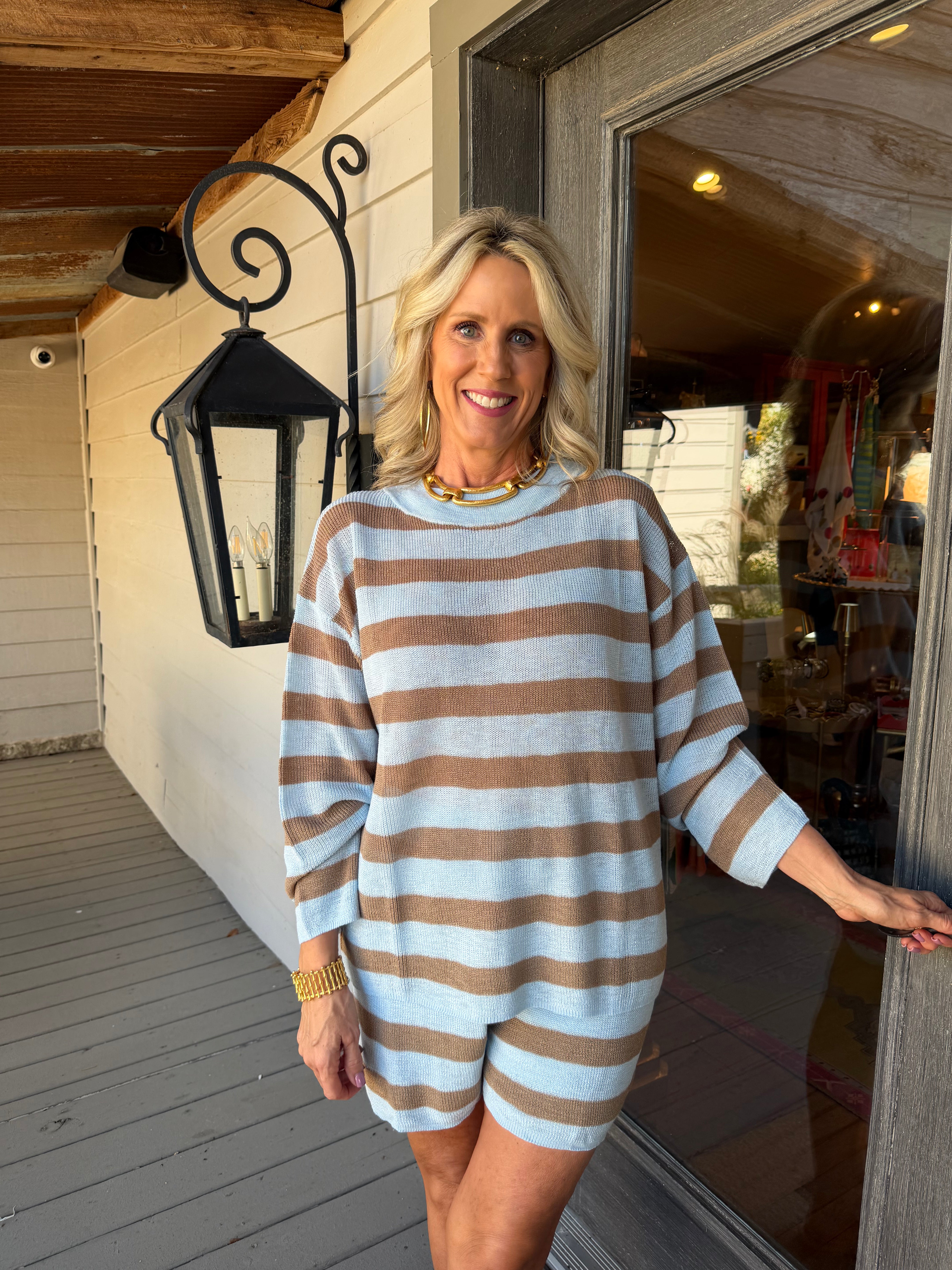 A smiling blonde woman stands on a porch wearing a loose, long-sleeve striped romper with alternating light blue and brown horizontal bands. The photo is taken in natural daylight beside a white house exterior with a black wall lantern and glass door.
