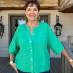 A woman wearing a bright green short-sleeve button-front cardigan and blue jeans, standing on a wooden deck in front of a house with beige siding and black lanterns.
