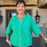 A woman wearing a bright green short-sleeve button-front cardigan and blue jeans, standing on a wooden deck in front of a house with beige siding and black lanterns.