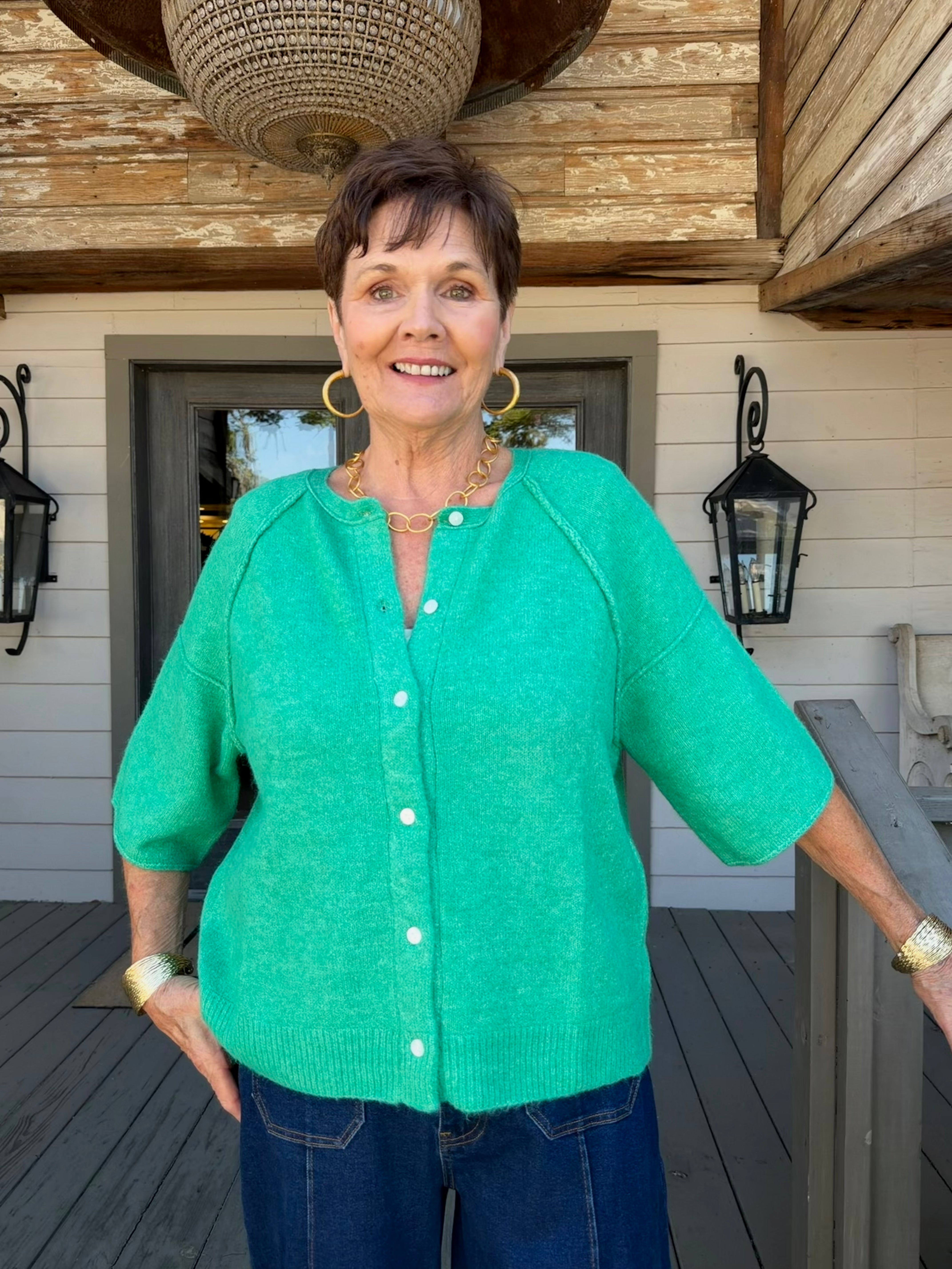A woman wearing a bright green short-sleeve button-front cardigan and blue jeans, standing on a wooden deck in front of a house with beige siding and black lanterns.