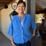A smiling woman stands on a porch wearing a bright blue knit cardigan with white buttons, dark blue jeans, and gold jewelry. The background shows a white wall, wooden ceiling beams, a black wall lantern, and a glass door.