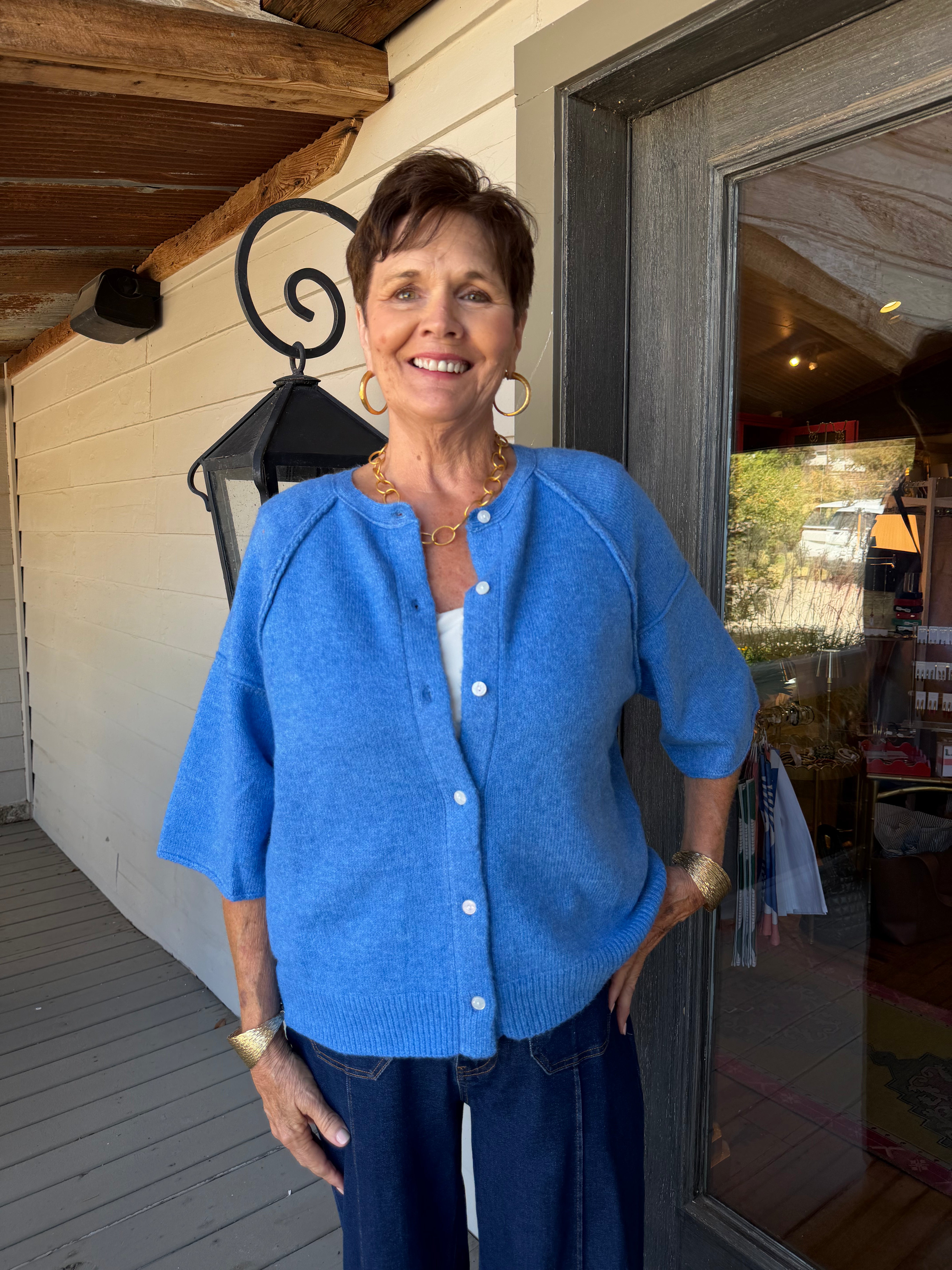 A smiling woman stands on a porch wearing a bright blue knit cardigan with white buttons, dark blue jeans, and gold jewelry. The background shows a white wall, wooden ceiling beams, a black wall lantern, and a glass door.