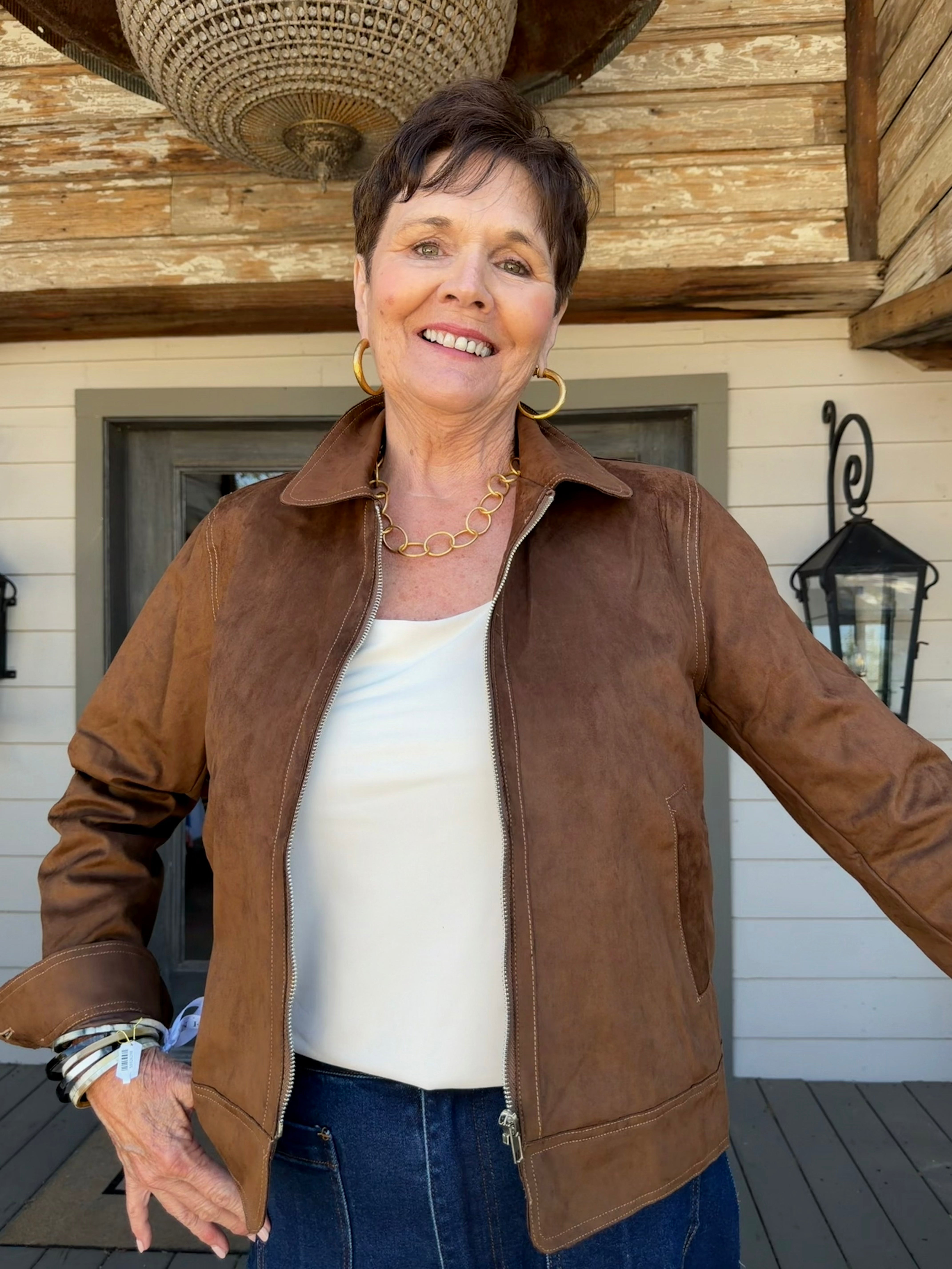 A smiling woman wearing a brown suede-like jacket over a white top and blue jeans, standing on a porch in front of a rustic wooden building.