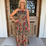 A woman stands in front of a rustic wooden door wearing a strapless midi dress with a beige base and a red, blue, and green floral print. She is accessorized with a gold necklace and bracelet.