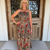A woman stands in front of a rustic wooden door wearing a strapless midi dress with a beige base and a red, blue, and green floral print. She is accessorized with a gold necklace and bracelet.