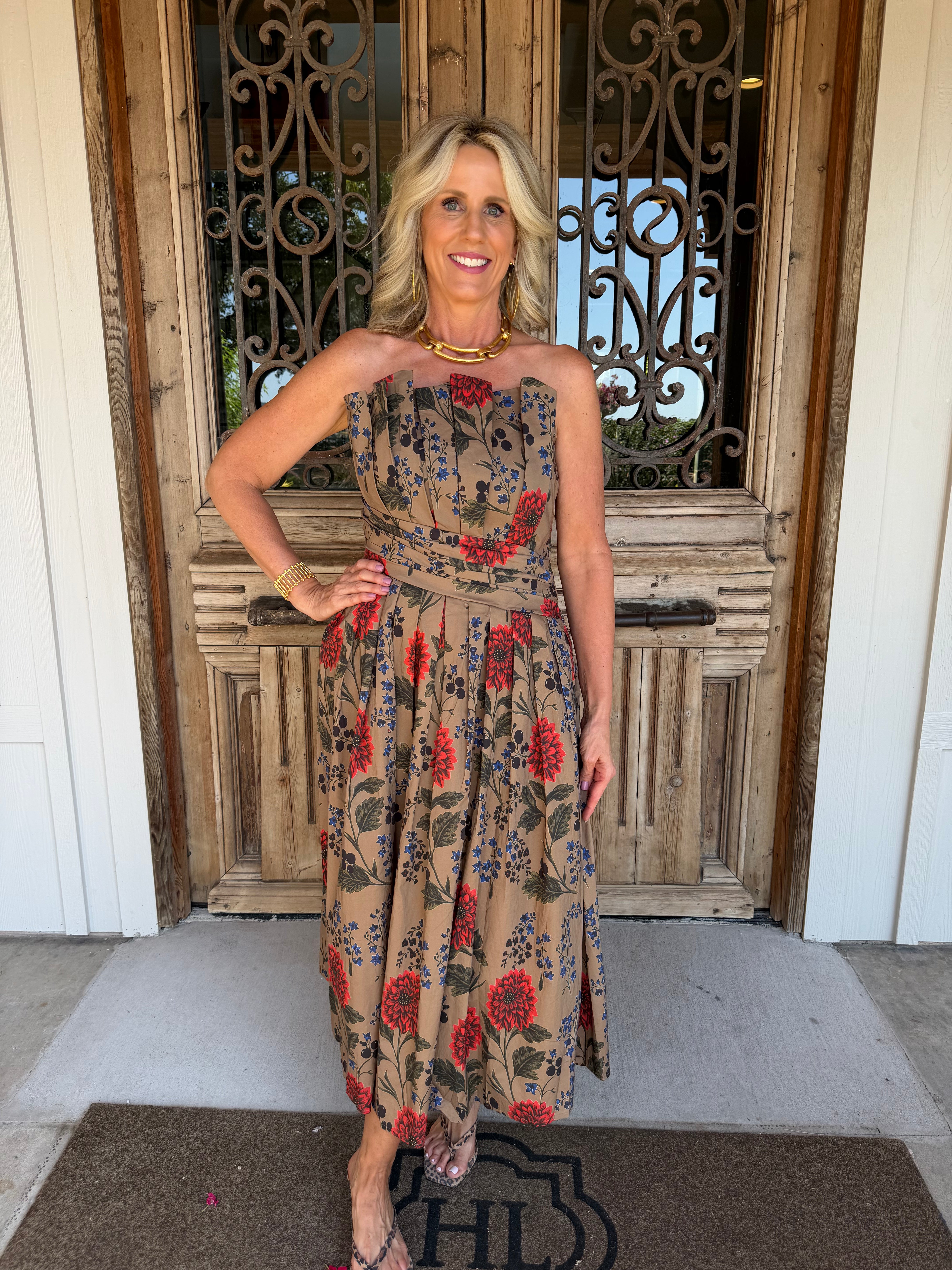A woman stands in front of a rustic wooden door wearing a strapless midi dress with a beige base and a red, blue, and green floral print. She is accessorized with a gold necklace and bracelet.