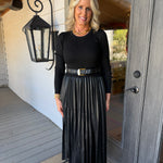 A woman stands on a porch wearing a black long-sleeve top and a black pleated midi skirt with a black belt featuring a gold buckle.