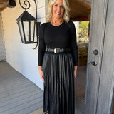 A woman stands on a porch wearing a black long-sleeve top and a black pleated midi skirt with a black belt featuring a gold buckle.