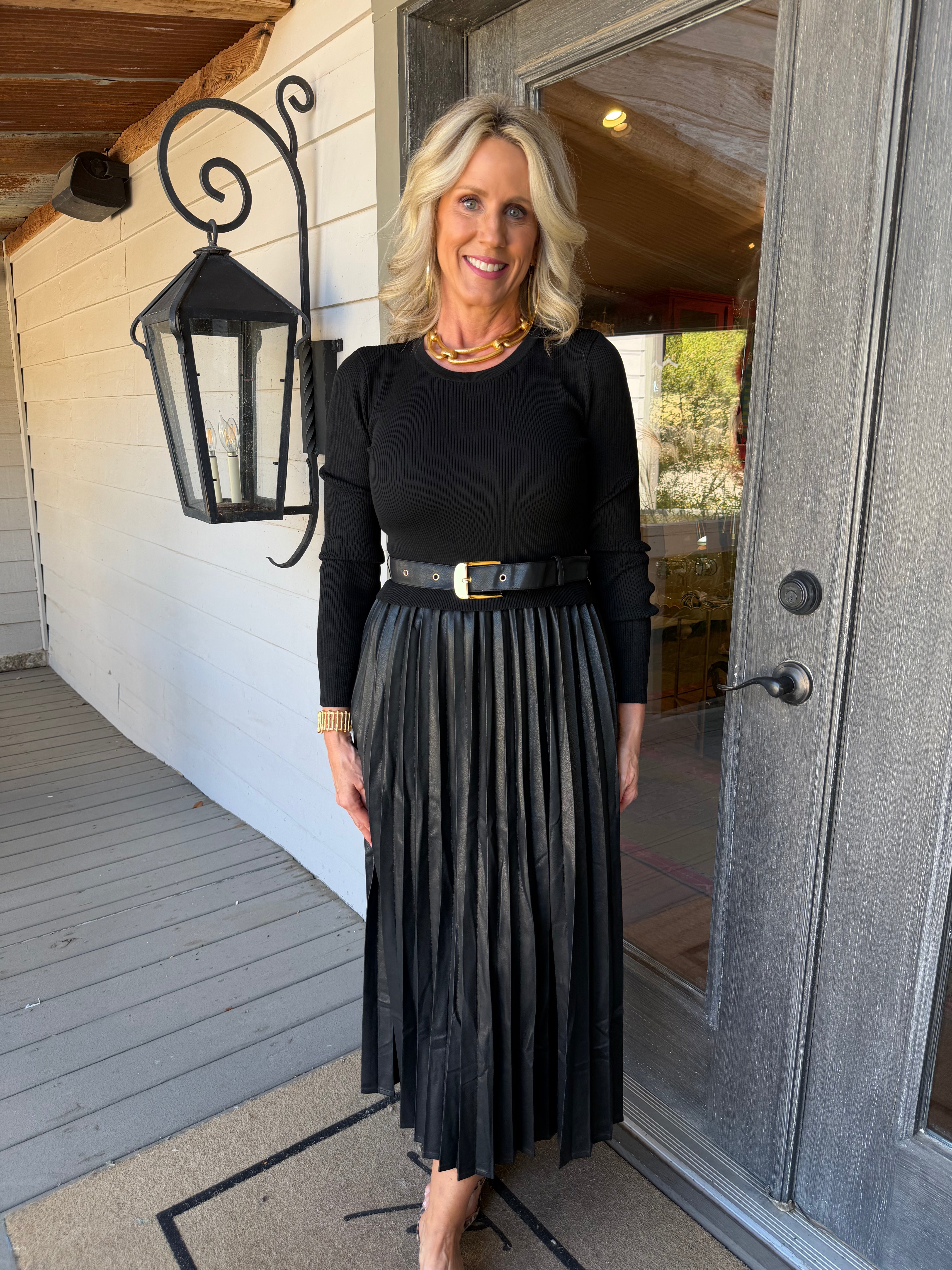 A woman stands on a porch wearing a black long-sleeve top and a black pleated midi skirt with a black belt featuring a gold buckle.