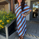 A woman stands on a wooden porch wearing a loose, knee-length dress with bold diagonal stripes in light blue and brown. The dress has a draped, kaftan-style silhouette with short wide sleeves.