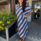 A woman stands on a wooden porch wearing a loose, knee-length dress with bold diagonal stripes in light blue and brown. The dress has a draped, kaftan-style silhouette with short wide sleeves.