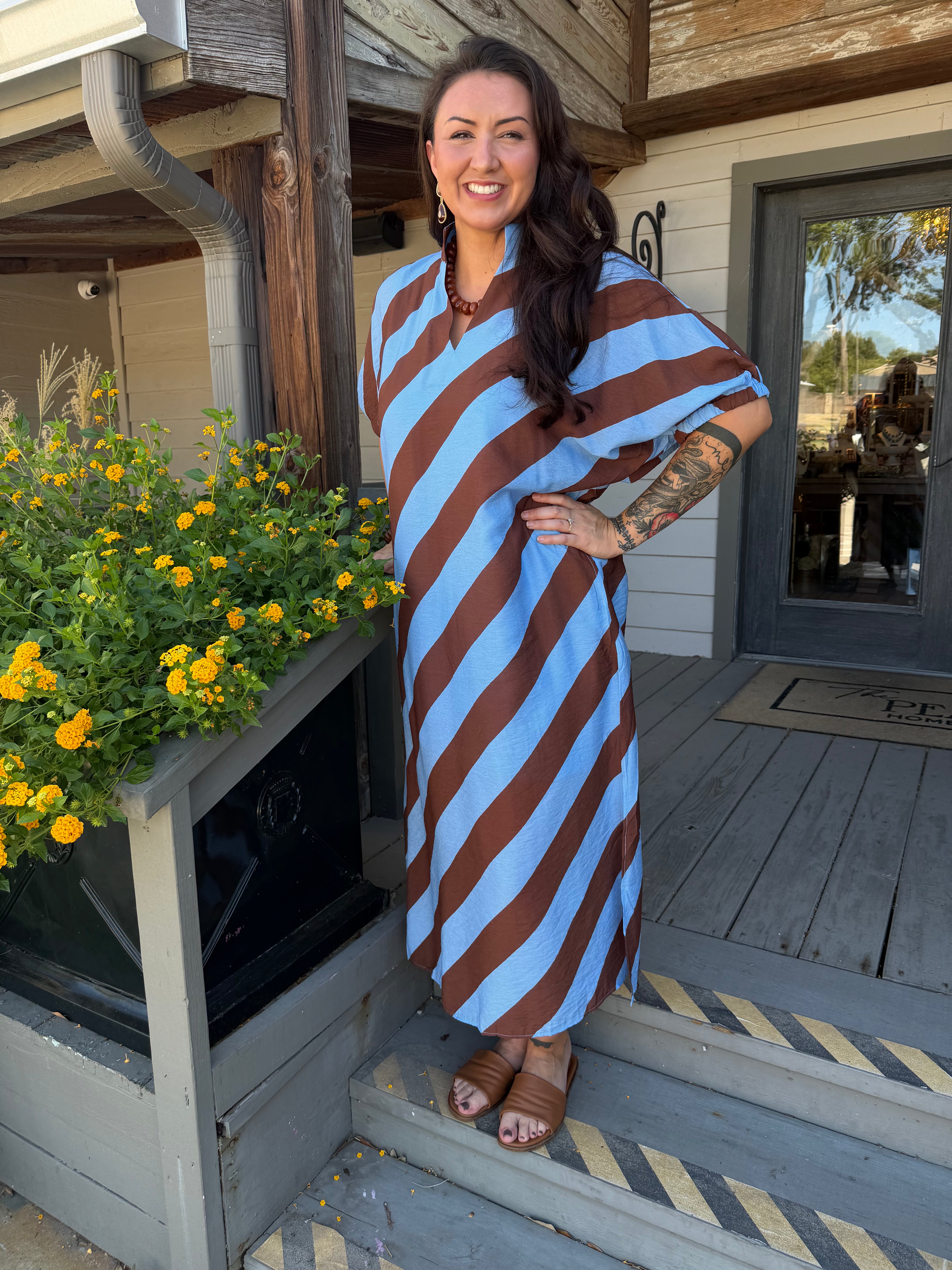 A woman stands on a wooden porch wearing a loose, knee-length dress with bold diagonal stripes in light blue and brown. The dress has a draped, kaftan-style silhouette with short wide sleeves.