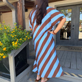 A woman wearing a brown and light blue striped dress stands on wooden porch steps facing a rustic wooden building with a door and yellow flowers in the foreground.