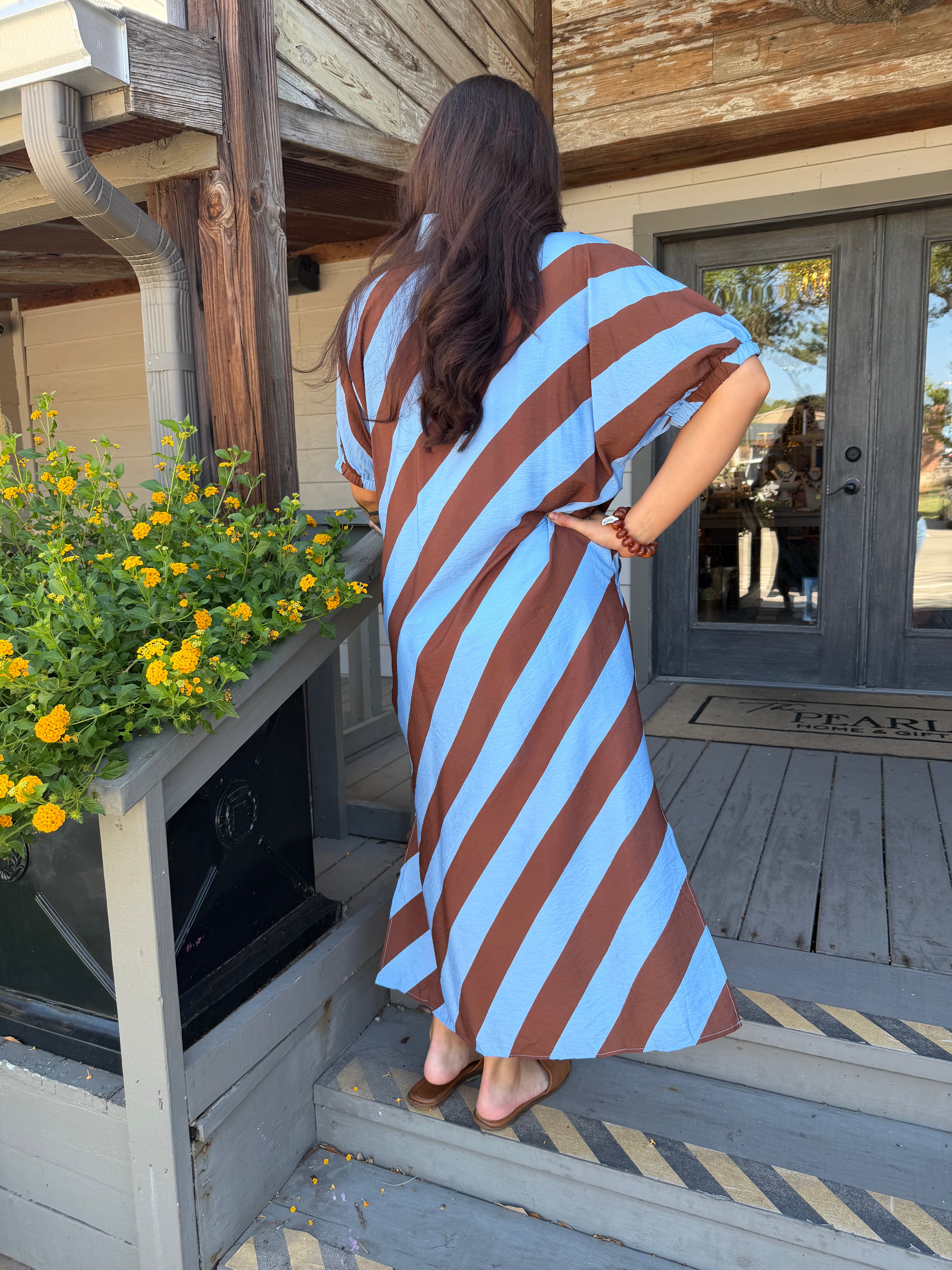 A woman wearing a brown and light blue striped dress stands on wooden porch steps facing a rustic wooden building with a door and yellow flowers in the foreground.
