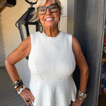 An older woman with short blonde hair and glasses is smiling while wearing a sleeveless white cable-knit top and patterned pants, standing on a porch next to a dark gray column.