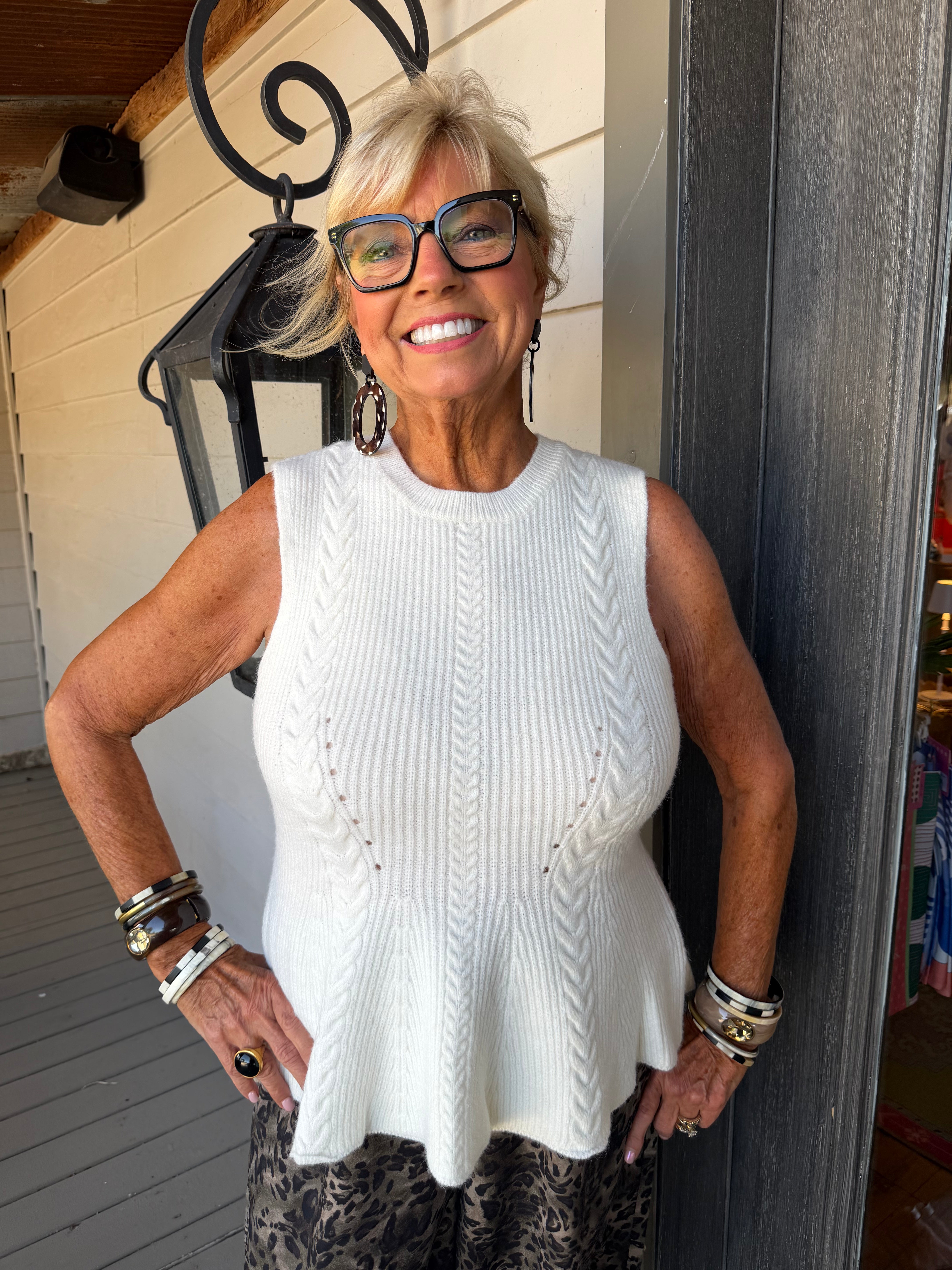 An older woman with short blonde hair and glasses is smiling while wearing a sleeveless white cable-knit top and patterned pants, standing on a porch next to a dark gray column.