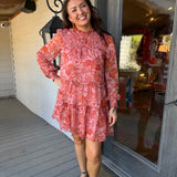 A woman stands on a porch wearing a long-sleeve ruffled mini dress with a coral and pink floral pattern, paired with brown slide sandals.