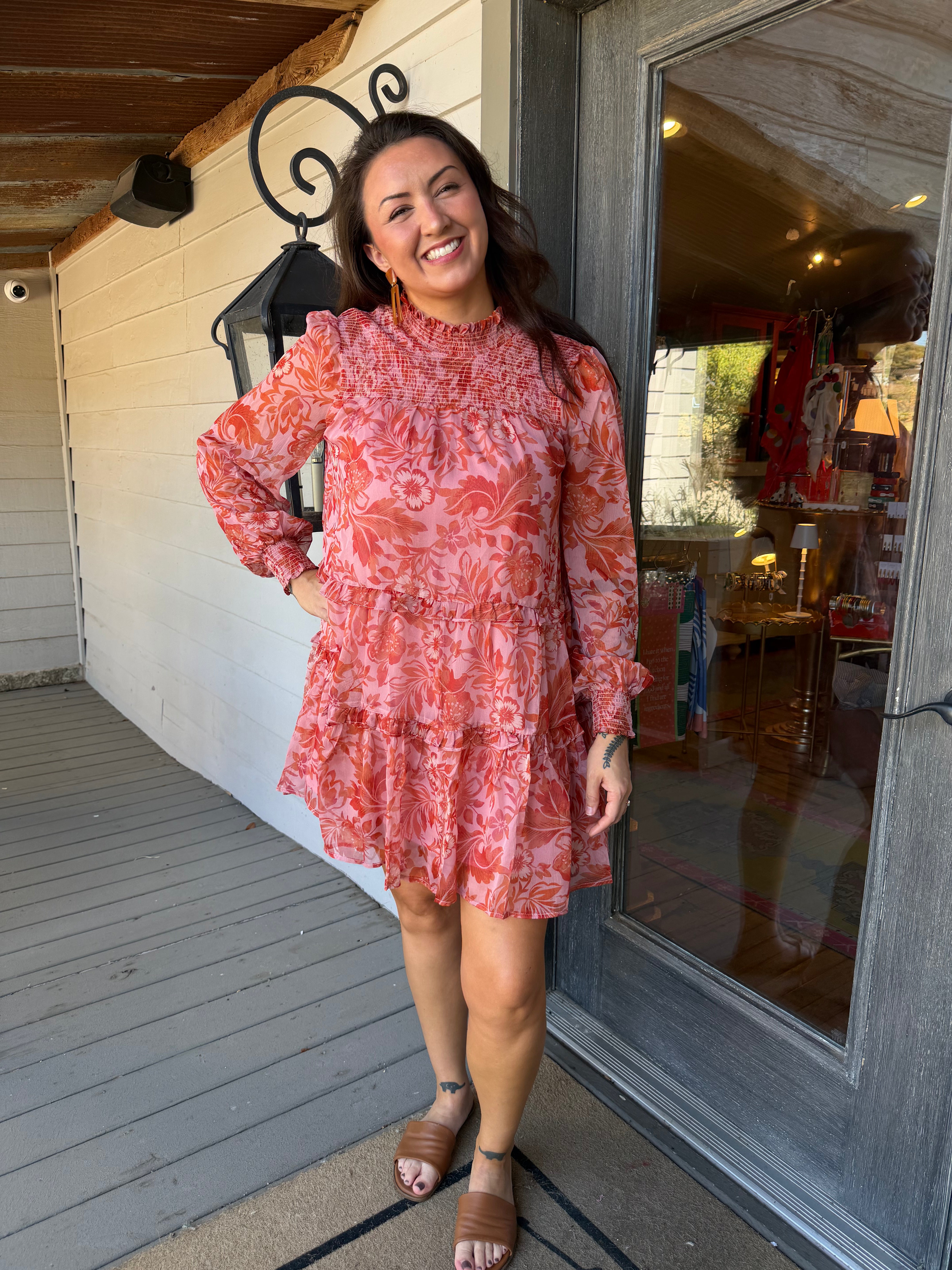 A woman stands on a porch wearing a long-sleeve ruffled mini dress with a coral and pink floral pattern, paired with brown slide sandals.