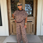 A woman wearing a brown and beige leopard-print lounge set stands on a front porch in front of a rustic wooden door.