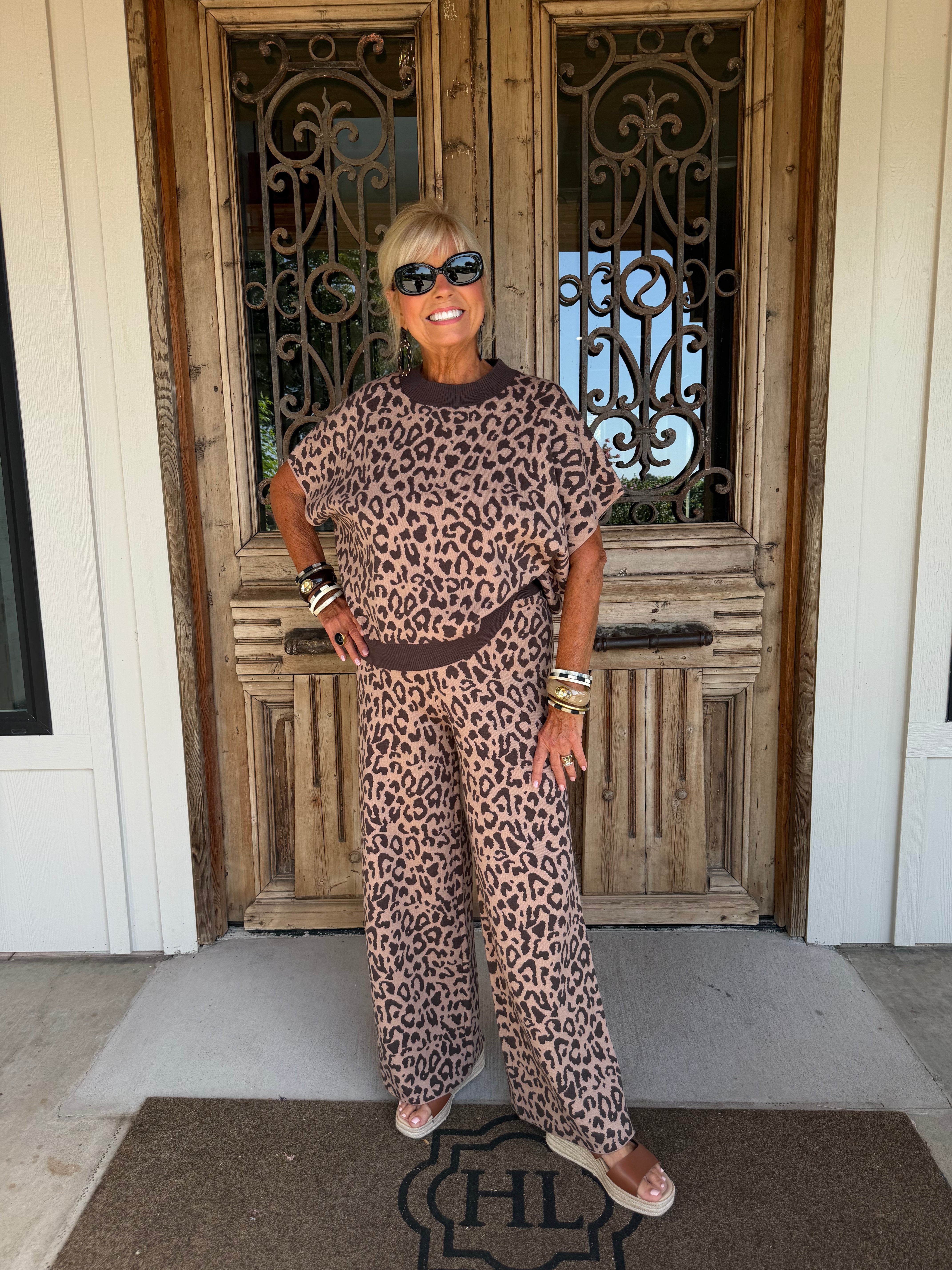 A woman wearing a brown and beige leopard-print lounge set stands on a front porch in front of a rustic wooden door.