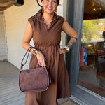 A woman stands on a porch wearing a brown sleeveless midi dress and holding a matching brown suede-look handbag. She is wearing large brown statement earrings and bracelets, with a glass door and white siding behind her.