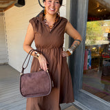 A woman stands on a porch wearing a brown sleeveless midi dress and holding a matching brown suede-look handbag. She is wearing large brown statement earrings and bracelets, with a glass door and white siding behind her.