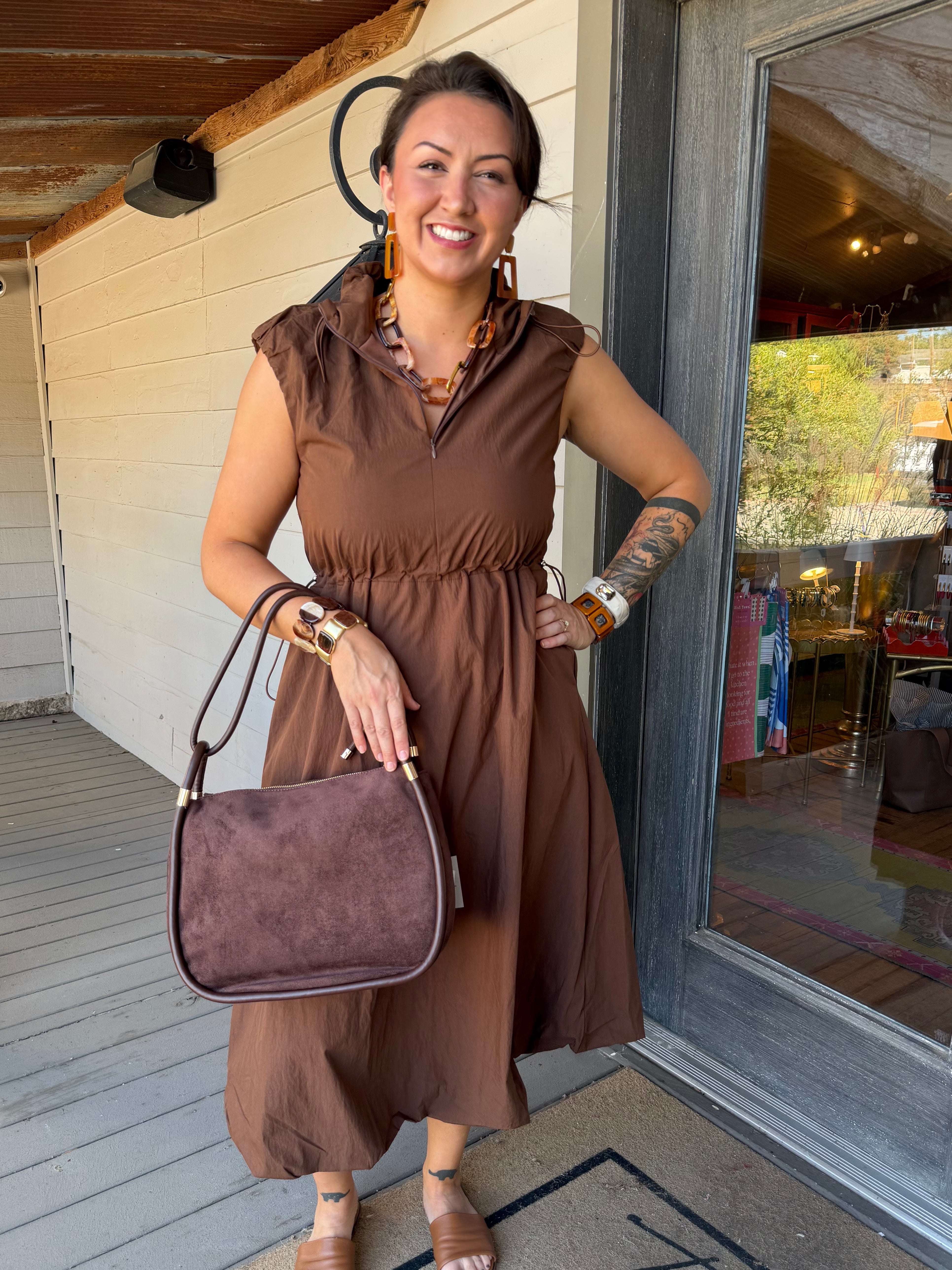 A woman stands on a porch wearing a brown sleeveless midi dress and holding a matching brown suede-look handbag. She is wearing large brown statement earrings and bracelets, with a glass door and white siding behind her.