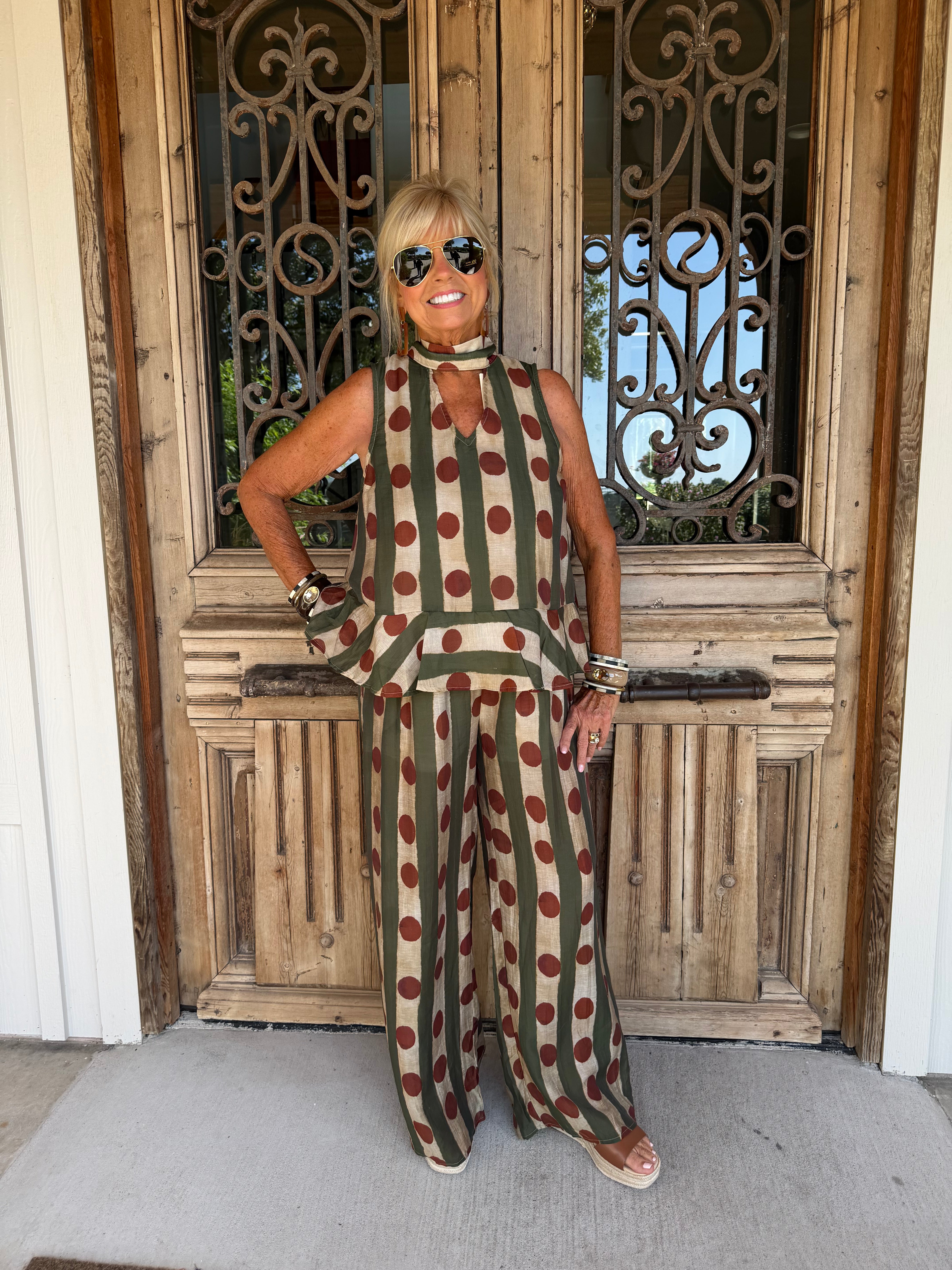 A woman modeling a sleeveless olive green and beige polka dot jumpsuit with a matching belt, standing in front of a rustic wooden door with decorative metal grates.
