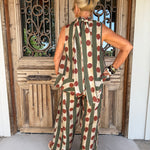 A woman viewed from behind wearing a sleeveless jumpsuit with vertical olive green and beige stripes and large red polka dots, standing in front of a rustic wooden door.