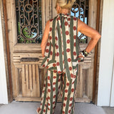 A woman viewed from behind wearing a sleeveless jumpsuit with vertical olive green and beige stripes and large red polka dots, standing in front of a rustic wooden door.