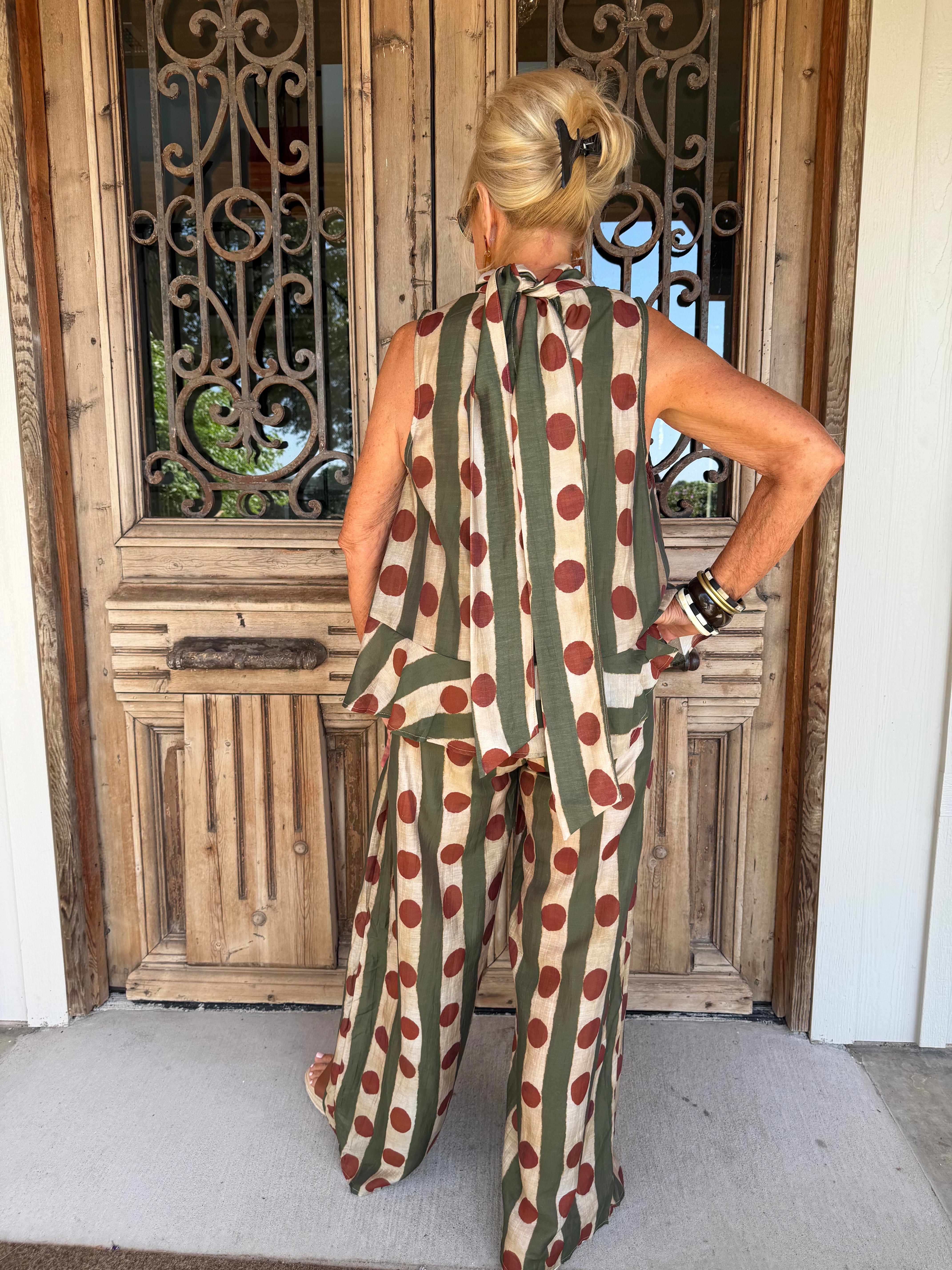 A woman viewed from behind wearing a sleeveless jumpsuit with vertical olive green and beige stripes and large red polka dots, standing in front of a rustic wooden door.
