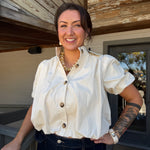 A woman smiling on a wooden porch, wearing a cream puff-sleeve button-up shirt and dark blue jeans.