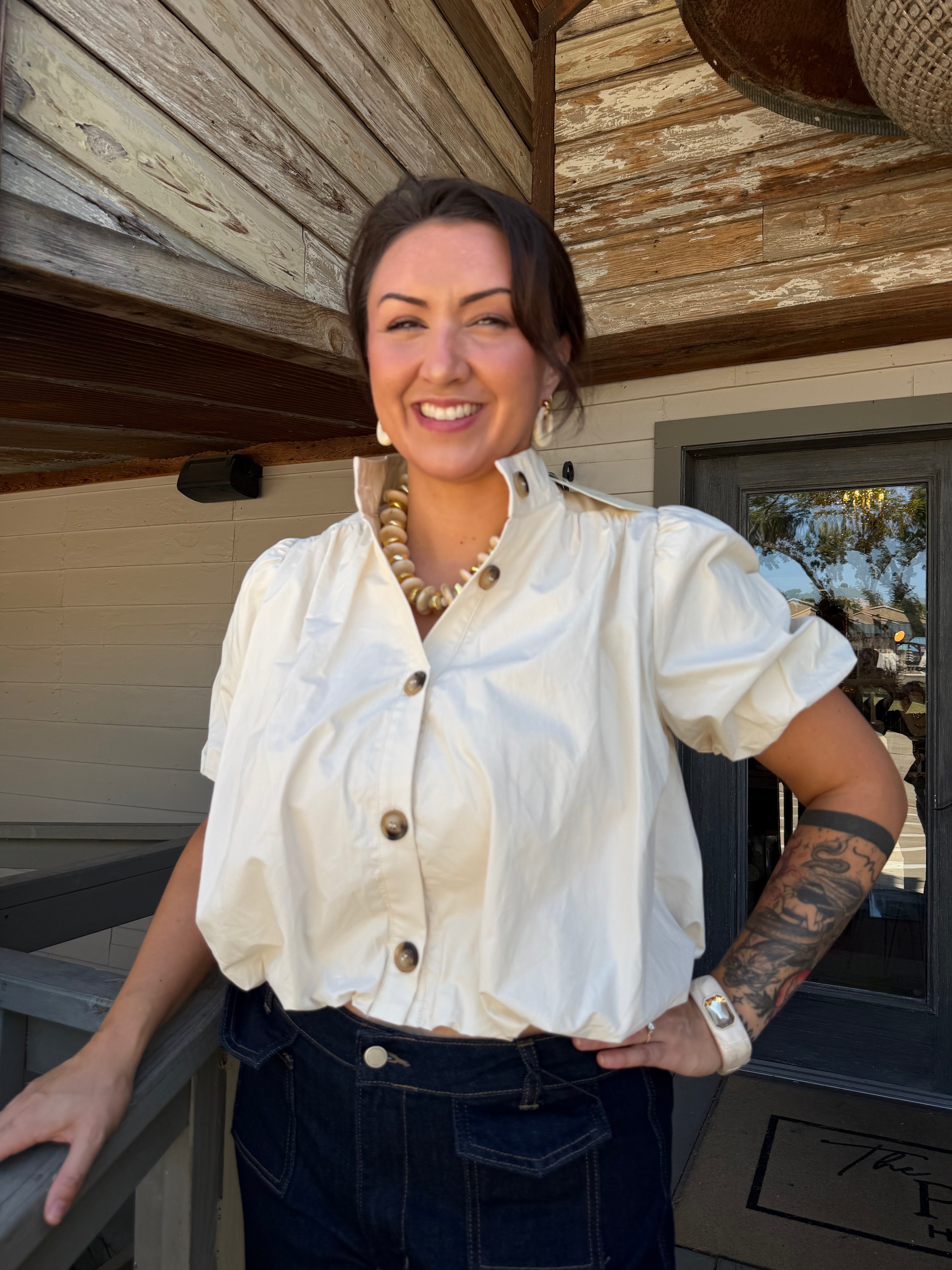 A woman smiling on a wooden porch, wearing a cream puff-sleeve button-up shirt and dark blue jeans.