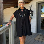 A woman with short gray hair and glasses stands on a wooden porch wearing a black short-sleeve button-front dress with a relaxed fit. She is accessorized with a chunky black necklace featuring gold and blue links, a black beaded collar, and a watch on her wrist.