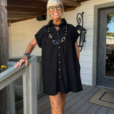 A woman with short gray hair and glasses stands on a wooden porch wearing a black short-sleeve button-front dress with a relaxed fit. She is accessorized with a chunky black necklace featuring gold and blue links, a black beaded collar, and a watch on her wrist.