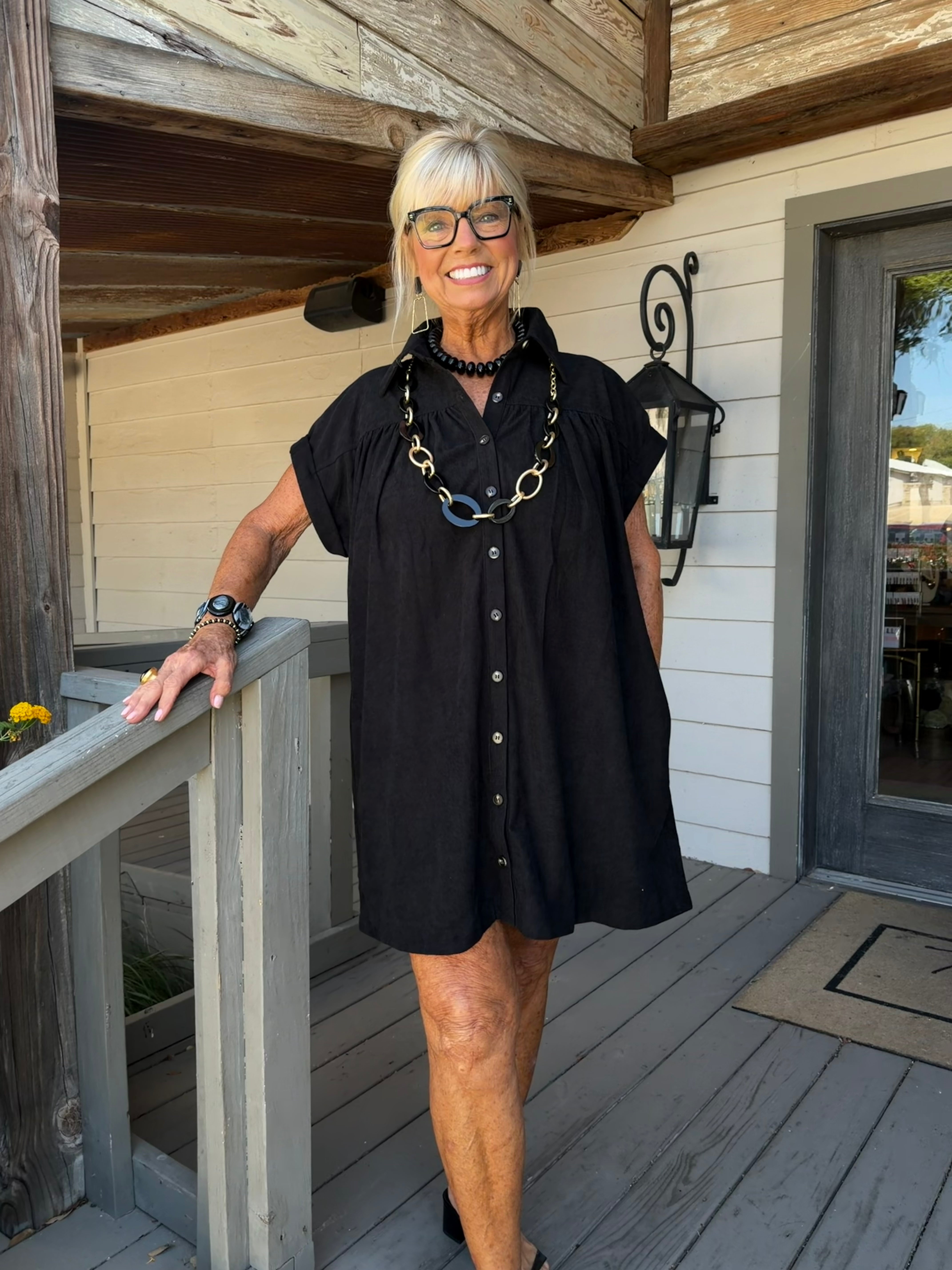 A woman with short gray hair and glasses stands on a wooden porch wearing a black short-sleeve button-front dress with a relaxed fit. She is accessorized with a chunky black necklace featuring gold and blue links, a black beaded collar, and a watch on her wrist.