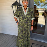 An older woman with short blonde hair and glasses stands on a porch wearing a short-sleeve leopard-print maxi dress with a V-neck and light beige trim. The dress has an olive green, black, and tan animal pattern.