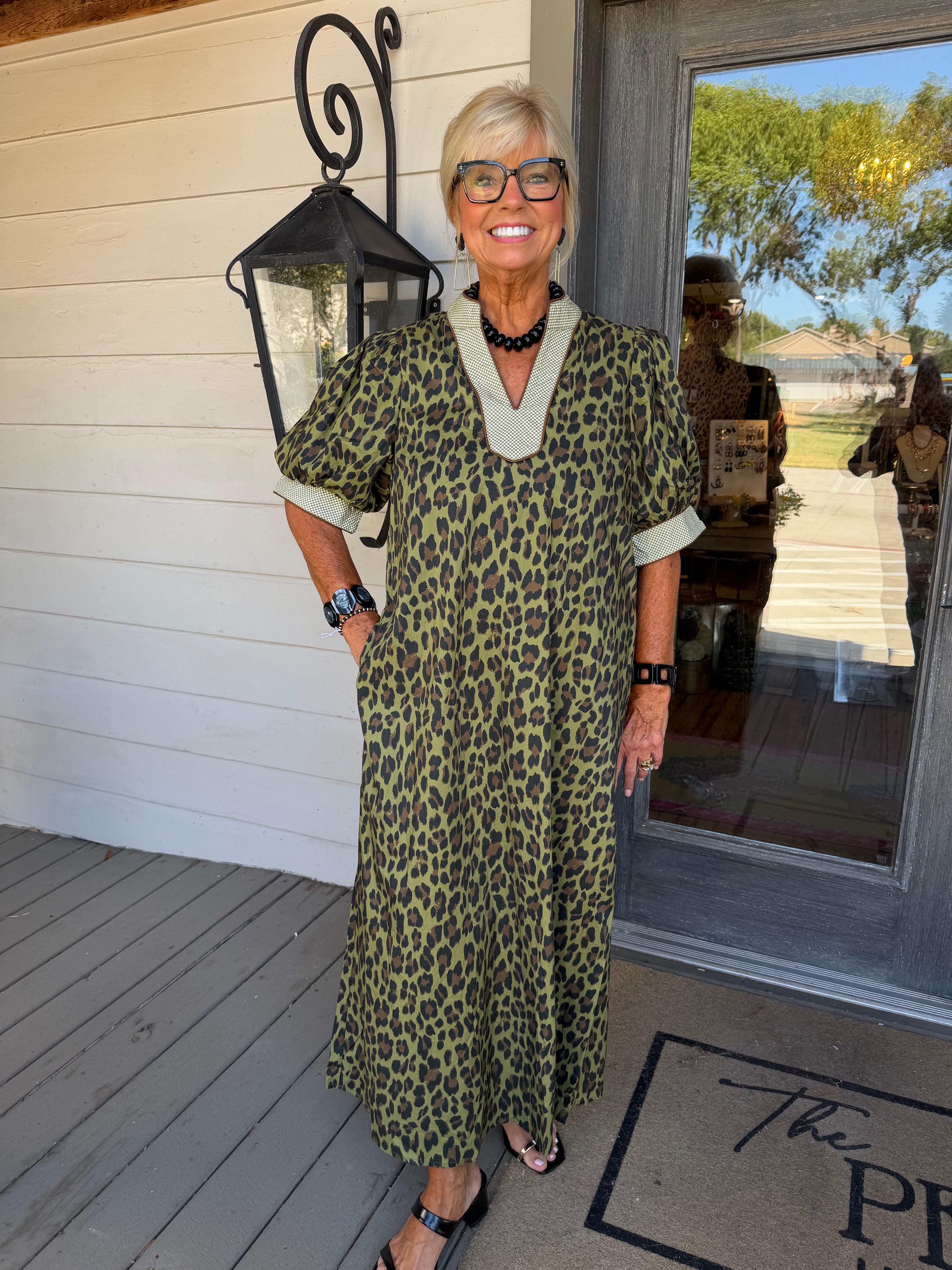 An older woman with short blonde hair and glasses stands on a porch wearing a short-sleeve leopard-print maxi dress with a V-neck and light beige trim. The dress has an olive green, black, and tan animal pattern.