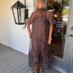 A woman wearing a sheer leopard-print midi dress stands on a porch in front of a white wood-paneled wall and glass door.