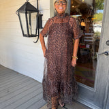 A woman wearing a sheer leopard-print midi dress stands on a porch in front of a white wood-paneled wall and glass door.