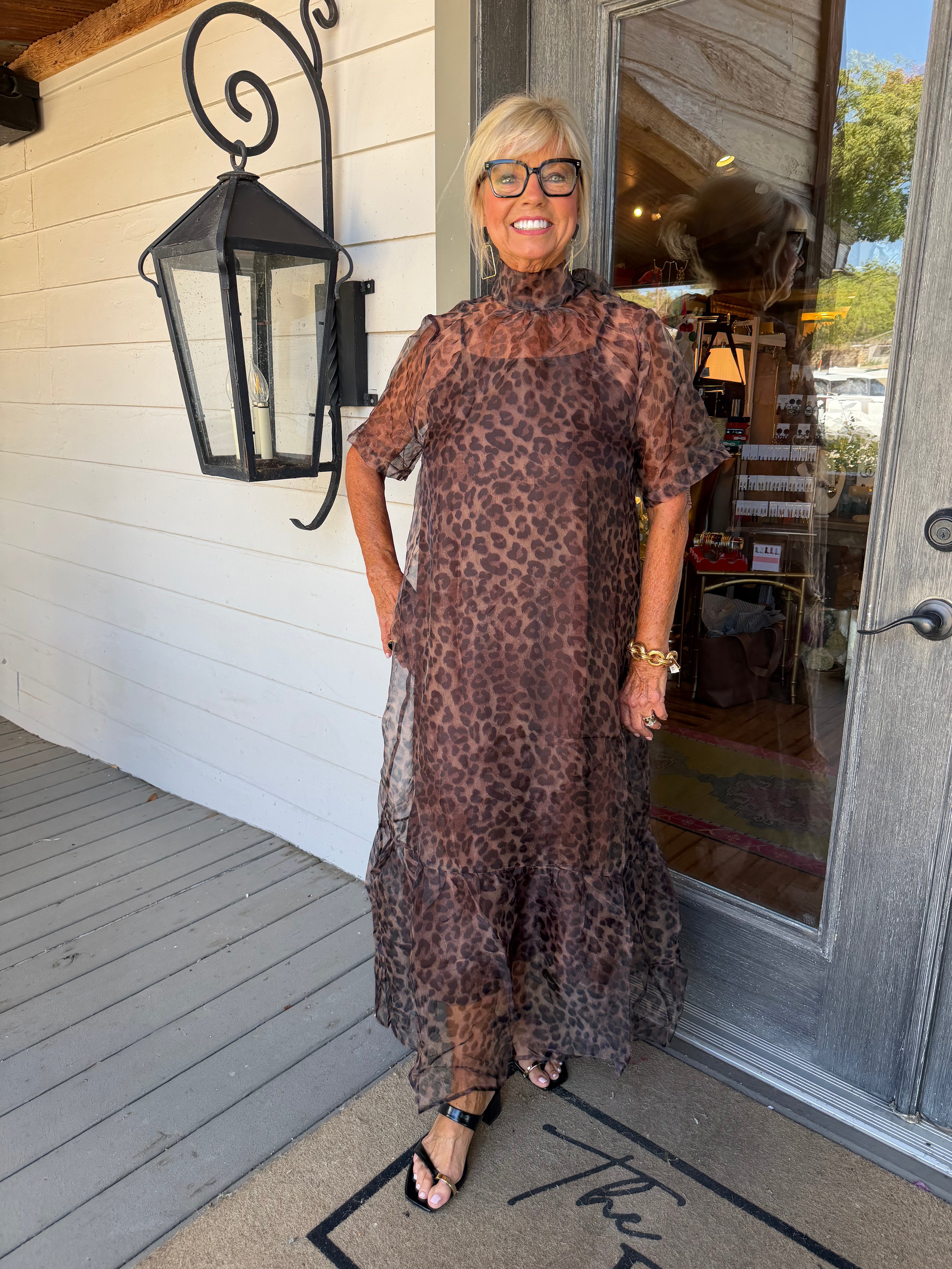 A woman wearing a sheer leopard-print midi dress stands on a porch in front of a white wood-paneled wall and glass door.