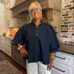 A smiling blonde woman wearing glasses, a dark navy blue puff-sleeve button-up blouse, and white pants, standing in a rustic kitchen with a large wood range hood and brick wall.