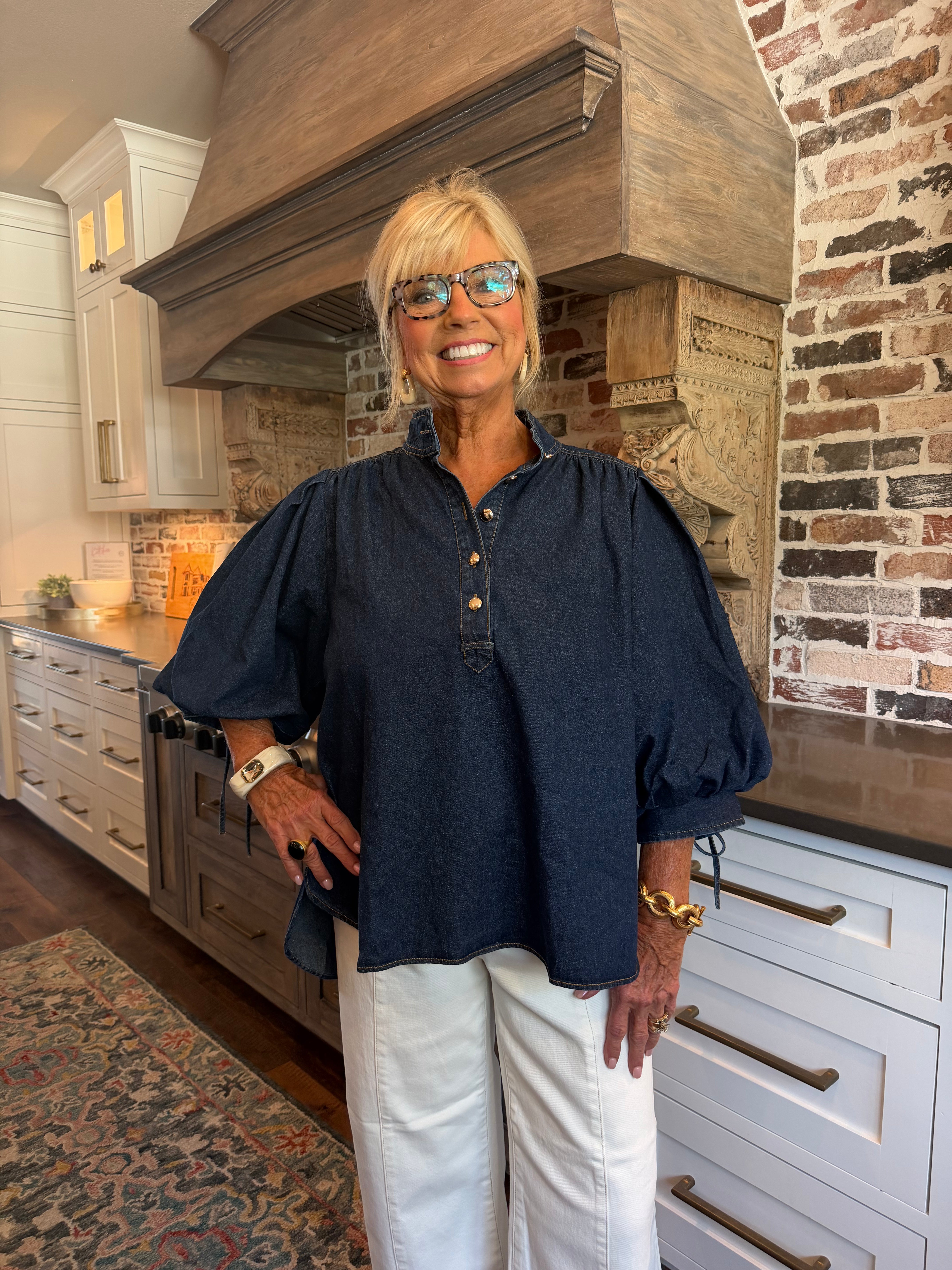 A smiling blonde woman wearing glasses, a dark navy blue puff-sleeve button-up blouse, and white pants, standing in a rustic kitchen with a large wood range hood and brick wall.