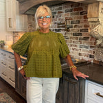 A smiling blonde woman wearing glasses, a textured olive green short-sleeve blouse, and white pants stands in a kitchen with a brick backsplash and dark wood cabinets.