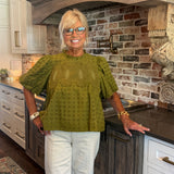 A smiling blonde woman wearing glasses, a textured olive green short-sleeve blouse, and white pants stands in a kitchen with a brick backsplash and dark wood cabinets.