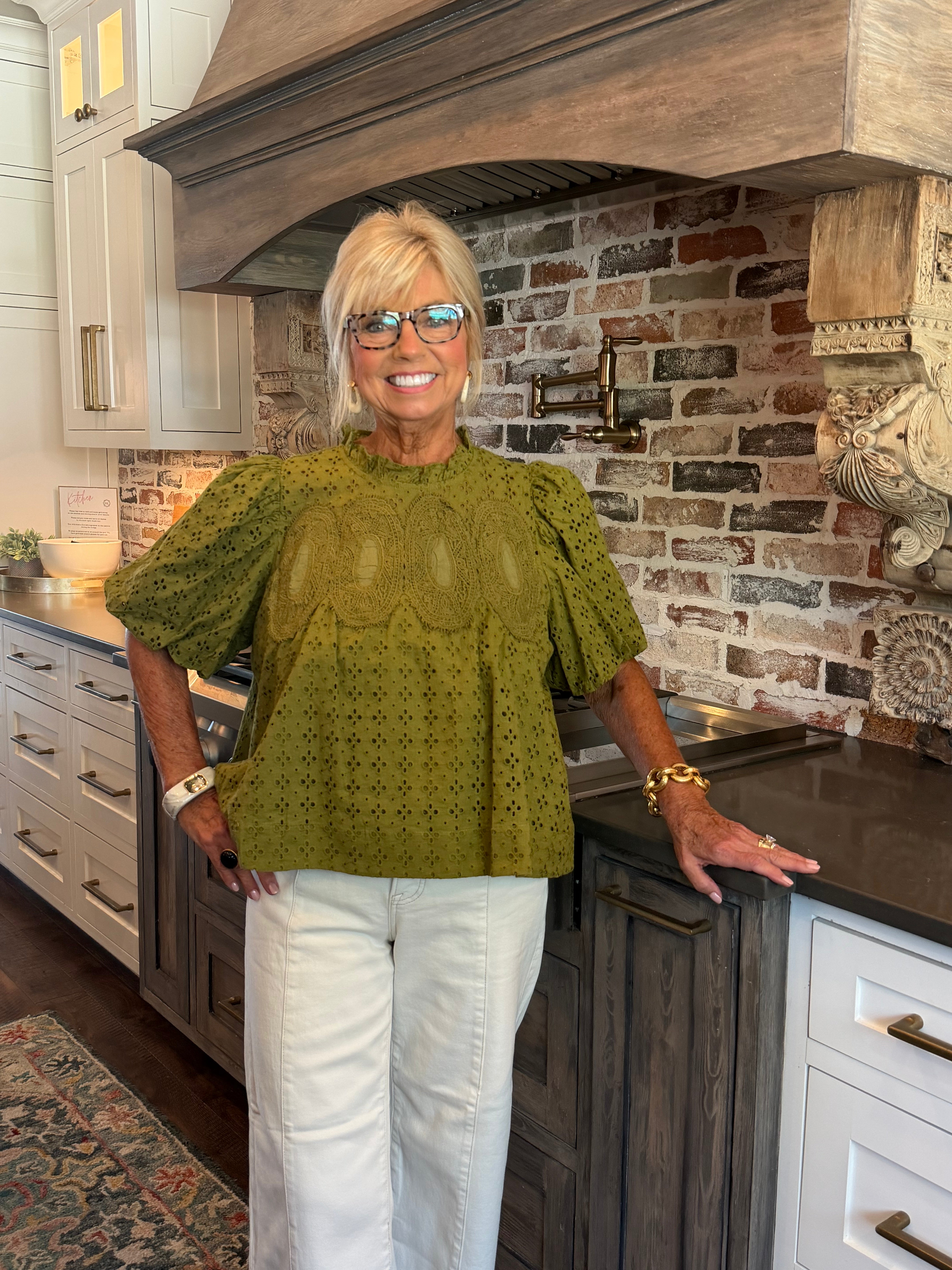 A smiling blonde woman wearing glasses, a textured olive green short-sleeve blouse, and white pants stands in a kitchen with a brick backsplash and dark wood cabinets.