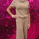 A woman modeling a beige knit short-sleeve top and matching wide-leg pants set, standing in front of a bright pink sequined wall.